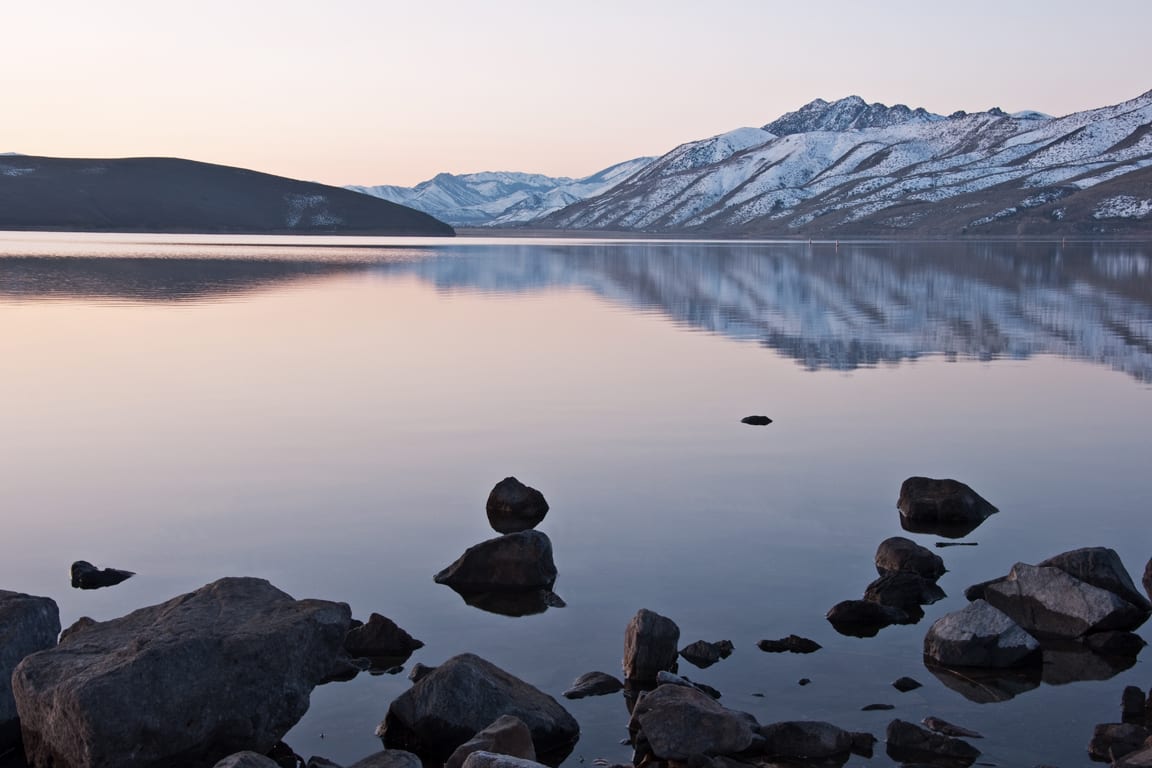 A lake with rocks in the foreground and mountains behind it.